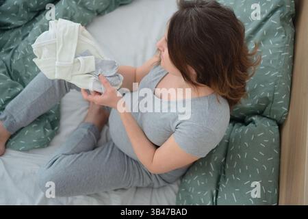 Eine glühende schwangere Frau sitzt im Kreuz, umgeben von Babykleidung und berührt sanft ihren Bauch. Ein ruhiger Moment der Liebe und Erwartung. Stockfoto