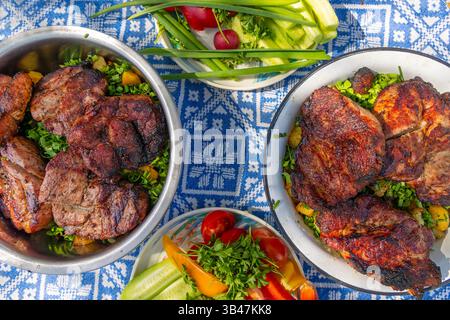 Gegrilltes Fleisch mit frischem Gemüse auf dem Tisch im Freien. Blick von oben auf gegrillte Schweinefleisch-Steaks, serviert mit Kräutern, gerösteten Kartoffeln und frischem Gartengemüse Stockfoto