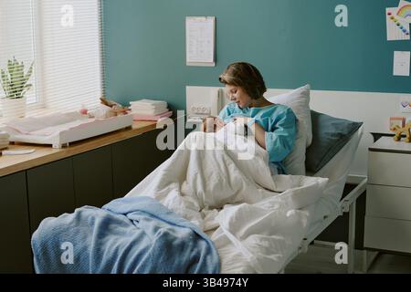 Krankenschwester, die das Neugeborene im Krankenhausbett hält, umgeben von klinischen Geräten und Babypflegeprodukten in einem gut organisierten Raum mit sanfter Beleuchtung und einer angenehmen Atmosphäre Stockfoto