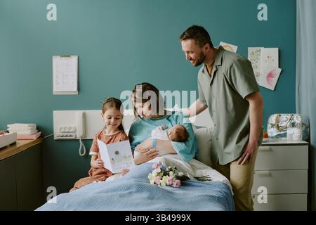 Glückliche Familie begrüßt neues Baby im Krankenhauszimmer mit Mutter, die Neugeborenes hält, Vater und Tochter in der Nähe sitzen, ein Buch lesen und den Moment zusammen genießen Stockfoto