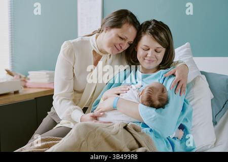 Die neue Mutter nimmt das Neugeborene auf, während Großmutter liebevoll zusieht. Zärtliche Bindungen der Familie, die sich zeigen, wenn Generationen besondere Momente teilen Stockfoto