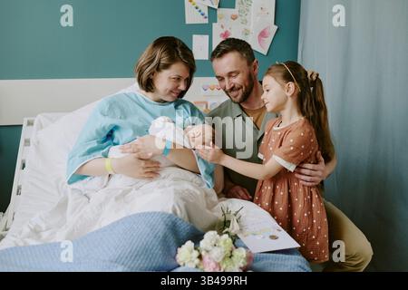 Glückliche Familie begrüßt Neugeborenes im Krankenhauszimmer mit Mutter, Vater und Tochter. Ein freudiger Moment, der festgehalten wird, als die Schwester sanft die Hand der Geschwister berührt Stockfoto
