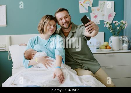 Glückliches Paar im Krankenhauszimmer, das Selfie macht, während es das Neugeborene hält. Hintergrund mit Plakaten, Pflanzen und persönlichen Gegenständen, die den intimen und fröhlichen Familienmoment unterstreichen Stockfoto
