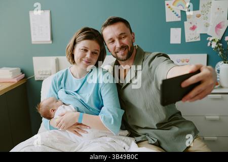 Die Familie lächelt, während sie Selfie mit dem Neugeborenen im Krankenhauszimmer macht und einen fröhlichen und intimen Moment während des postpartalen Krankenhausaufenthalts festnimmt Stockfoto