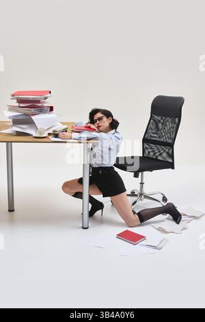 Porträt einer jungen Frau, die erschöpft auf dem Arm unter einem überladenen Schreibtisch in Büroumgebung vor weißem Hintergrund aussieht. Stockfoto