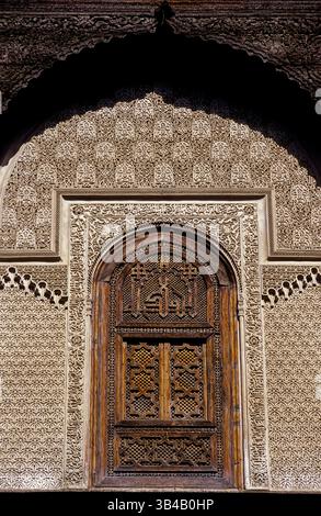 Geschnitztes Zedernholz, umgeben von koranischer Kalligraphie und kunstvollen Stuckmotiven im Innenhof der mittelalterlichen „Schule der Parfümeure“-Schule, der Al-Attarine Madrasa in Fes el-Bali Medina in Fes oder Fes in Nordmarokko. Die Madrasa, erbaut 1323-25, ist bekannt für ihre hochwertige Architektur und Dekoration. Stockfoto