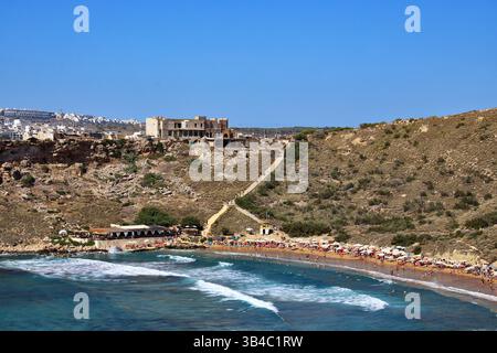 Geschäftiger Ramla Bay Strand in Gozo, Malta, mit goldenem Sand, Sonnenanbetern, Wellen, und ein malerischer Pfad führt zu einem verlassenen Steingebäude auf dem Hügel. Stockfoto