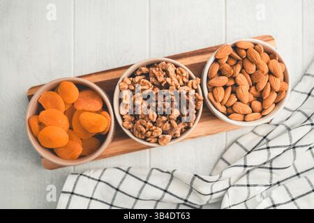 Drei Schüsseln mit gesunden Snacks, darunter getrocknete Aprikosen, Walnüsse und Mandeln, sind auf einem Holzbrett angeordnet und bieten eine nahrhafte und köstliche Leckerei Stockfoto