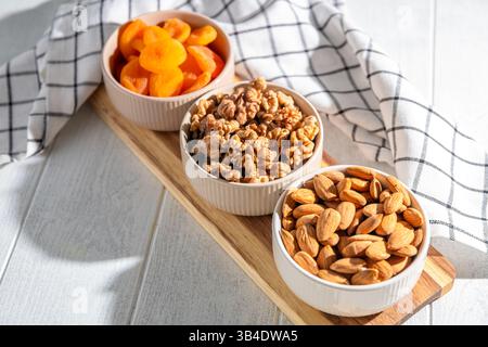 Drei Schüsseln mit gesunden Snacks, darunter getrocknete Aprikosen, Walnüsse und Mandeln, sind auf einem Holzbrett angeordnet und bieten eine nahrhafte und köstliche Leckerei Stockfoto