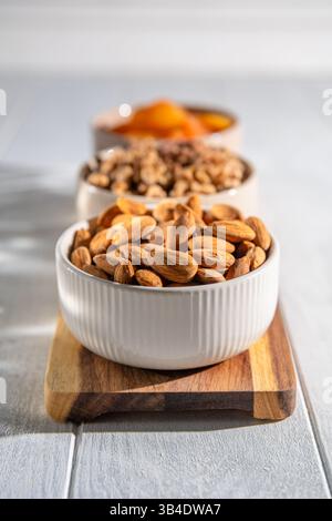 Drei Schüsseln mit gesunden Snacks, darunter getrocknete Aprikosen, Walnüsse und Mandeln, sind auf einem Holzbrett angeordnet und bieten eine nahrhafte und köstliche Leckerei Stockfoto