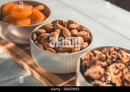 Drei Schüsseln mit gesunden Snacks, darunter getrocknete Aprikosen, Walnüsse und Mandeln, sind auf einem Holzbrett angeordnet und bieten eine nahrhafte und köstliche Leckerei Stockfoto