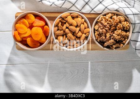 Drei Schüsseln mit gesunden Snacks, darunter getrocknete Aprikosen, Walnüsse und Mandeln, sind auf einem Holzbrett angeordnet und bieten eine nahrhafte und köstliche Leckerei Stockfoto
