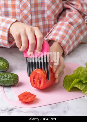 Kind, das mit der Zubereitung von Salat und dem Schneiden von Tomaten mit einem welligen Edelstahlmesser auf rosafarbenem Schneidebrett beschäftigt ist Stockfoto
