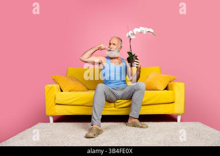 Stilvoller Seniorenmann, der auf einem gelben Sofa mit einer Blume posiert, vor einem leuchtend rosa Hintergrund in einer ungezwungenen Innenumgebung. Stockfoto