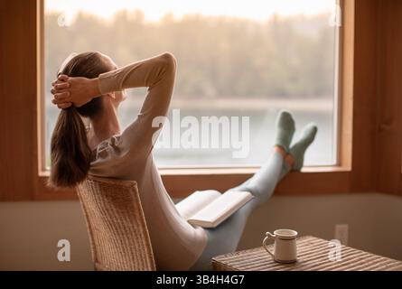 Person, die sich zu Hause beim Lesen eines Buches entspannt an einem gemütlichen Wintermorgen bei einer Tasse heißem Tee Kaffee fühlt Stockfoto
