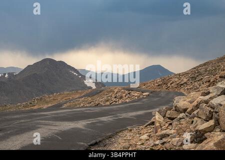 Eine kurvige Straße schlängelt sich durch raues Gelände, umgeben von felsigen Felsvorsprüngen und fernen Bergen. Dunkle Wolken erheben sich über ihnen und deuten auf ein mögliches Sto hin Stockfoto