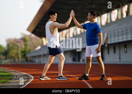 Zwei asiatische Männer stehen auf der Rennstrecke im Sportstadion und geben sich gegenseitig High Five. Sportlichkeit, Outdoor-Training oder Training. Stockfoto