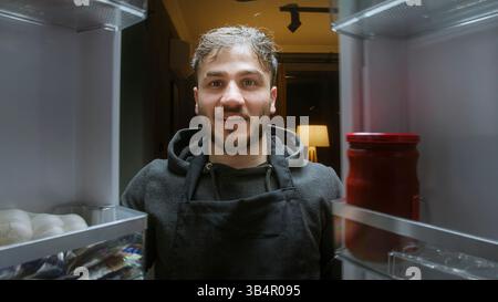 Ein lächelnder Mann, der nachts aus der Sicht des Kühlschranks ein Glas mit Essen im Kühlschrank findet Stockfoto