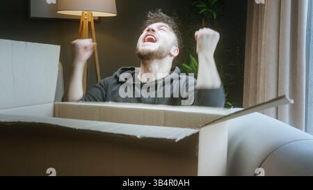Junger Mann, der ein Paket öffnet und überrascht reagiert, während er zu Hause sitzt Stockfoto