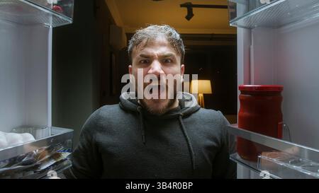 Ein wütender Mann, der mit leeren Händen im Kühlschrank aus der Sicht des Kühlschranks brüllt Stockfoto