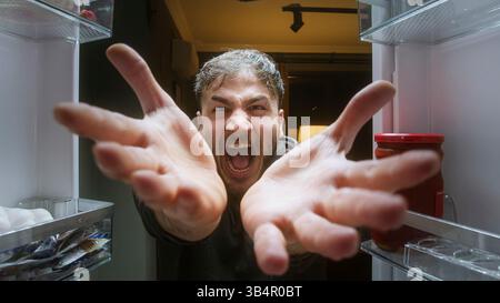 Wütender hungriger Mann, der mit leeren Händen im Kühlschrank aus der Sicht des Kühlschranks brüllt Stockfoto