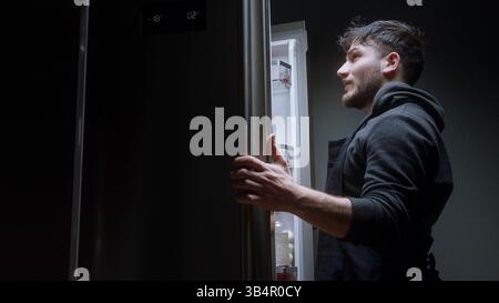 Frustrierter Mann, der nach Essen im Kühlschrank sucht und nicht findet, was er nachts will Stockfoto
