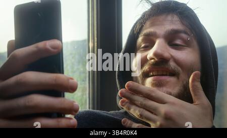 Ein Mann lächelt und fotografiert sich mit seinem Handy. Er trägt einen Hoodie und hat einen Bart Stockfoto