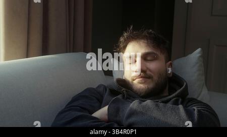 Entspannter junger Mann, der Musik genießt, mit geschlossenen Augen auf einer Couch im Sonnenlicht Stockfoto