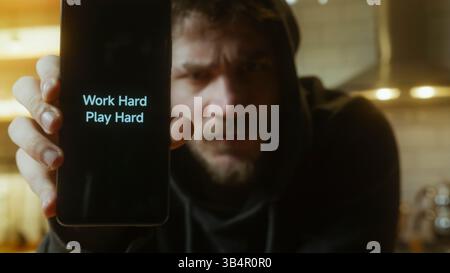 Motivierter junger Mann, der einen Telefonbildschirm mit Work Hard Play Hard Message in schwach beleuchtetem Raum zeigt Stockfoto