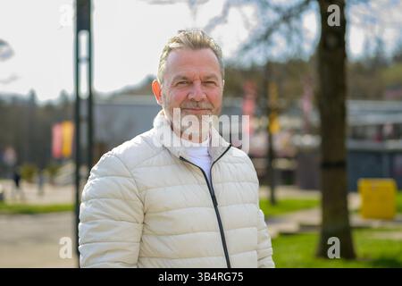 Ein fröhlicher Seniorenmann steht selbstbewusst in einem sonnendurchfluteten Park, trägt eine leichte Pufferjacke und ein lässiges weißes Hemd. Stockfoto