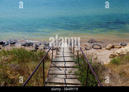 Alte Holztreppe mit rostigem Geländer, die zu einem verschmutzten Strand mit abgenutzten Reifen und klarem türkisfarbenem Wasser an einem sonnigen Tag hinabführt Stockfoto