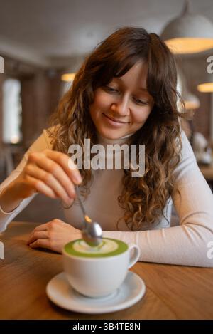 Junge Frau mit lockigem Haar, die ihren Matcha-Latte in einem gemütlichen Café umrührt, die warme Atmosphäre genießt und einen ruhigen Moment voller Entspannung und Komfort genießt Stockfoto