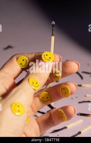 Ein verbrannter Streichhölzer in der Hand unter vielen Streichhölzern auf hellviolettem Hintergrund. Gelbe Emoticons, kreativ. Stockfoto