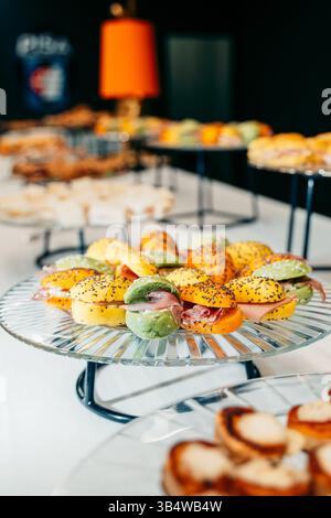 Verschiedene Gourmet-Sandwiches mit bunten Brötchen sind kunstvoll auf eleganten Glastabletts angeordnet. Perfekt für Catering, Empfänge und besondere Anlässe, Stockfoto