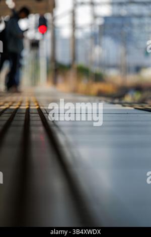 Tiefwinkelansicht des Bahnsteigsteigs mit taktilem Pflaster und Bokeh-Hintergrundfigur. Ein rot leuchtendes Signal erhöht die Erwartung einer wartenden Person. Stockfoto