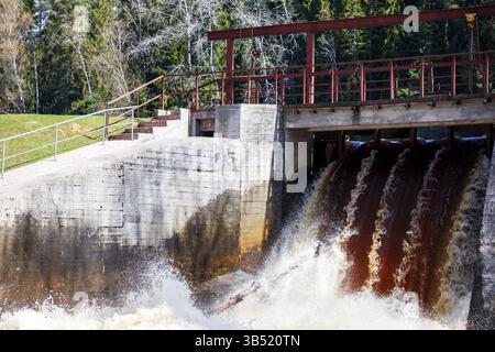 Ein kleiner Wasserfall oder Staudamm an einem Fluss, wo Wasser herabstürzt und Schaum bildet, und die Struktur besteht aus Beton und Metall mit roten Geländern. A für Stockfoto