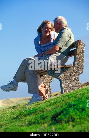Seniorenmann mit erwachsener Tochter sitzt draußen auf einer Bank am Hügel. Stockfoto