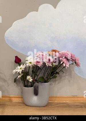 Das Bild zeigt eine Gruppe von Blumen, darunter rote Rosen, weiße Gänseblümchen und rosa Chrysanthemen, die in einer modernen U-förmigen, gerippten Keramikvase angeordnet sind. Th Stockfoto