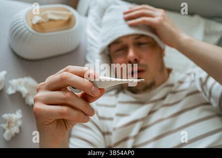 Kranker Mann mit Erkältung oder Grippe, liegend im Bett mit nassem Handtuch auf der Stirn, überprüft seine Körpertemperatur mit Thermometer, Taschentücher und benutzten Taschentüchern auf dem Bett Stockfoto