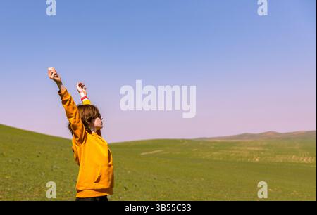 Ein Teenager, der bei Sonnenuntergang mit erhobenen Händen auf freiem Feld steht, digitale Entgiftung und Verbindung mit der Natur genießt. Entspannte Sommeratmosphäre und friedliche Atmosphäre Stockfoto