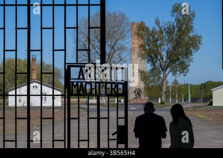 Oranienburg, Deutschland. April 2025. Das eiserne Tor am Eingang des Gefangenenlagers „Turm A“ mit der Aufschrift „Arbeit macht frei“ an der Gedenkstätte Sachsenhausen. Im Hintergrund befindet sich der rund 40 Meter hohe Obelisk, das zentrale Denkmal und Wahrzeichen der 1961 eröffneten DDR-Nationaldenkmal Sachsenhausen. Anlässlich des 80. Jahrestages der Befreiung bietet die Stiftung Brandenburgische Gedenkstätten ein abwechslungsreiches Programm an Ausstellungen, Veranstaltungen und pädagogischen Aktivitäten an. Quelle: Soeren Stache/dpa/Alamy Live News Stockfoto