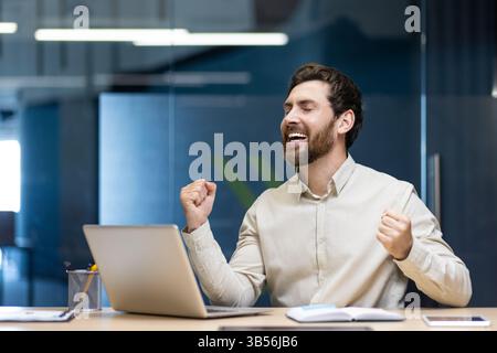 Ein glücklicher junger Geschäftsmann sitzt im Büro am Tisch und freut sich über den Erfolg und das Ergebnis und hebt die Hände hoch. Stockfoto