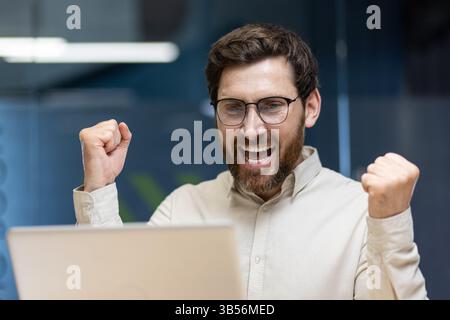 Nahaufnahme eines jungen glücklichen Mannes, der im Büro ist, auf den Laptop-Bildschirm blickt und glücklich ist, und mit seinen Händen eine Siegesgeste zeigt. Stockfoto