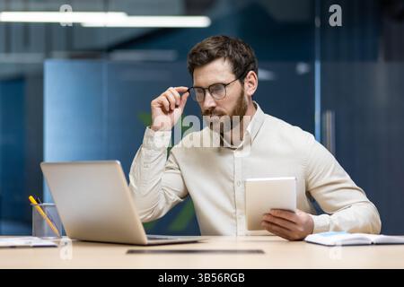 Foto eines jungen Geschäftsmannes, der am Arbeitsplatz im Büro sitzt, ein Tablet benutzt, eine Brille hält und überrascht auf den Laptop-Bildschirm blickt. Stockfoto