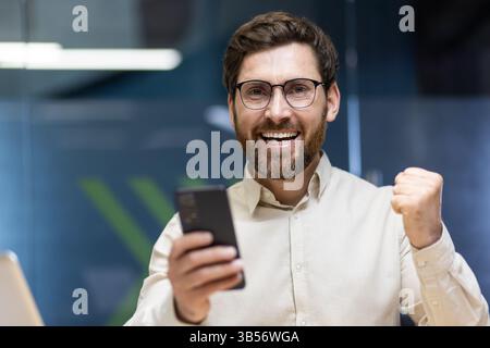 Nahporträt eines jungen glücklichen Mannes in Brille und hellem Hemd, der im Büro ist, sich über Erfolg und Sieg freut, ein Telefon in der Hand hält. Stockfoto