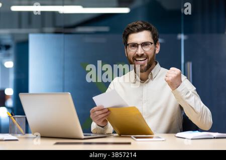 Nahporträt eines glücklichen jungen Mannes, der an einem Schreibtisch im Büro sitzt. Hält einen offenen Umschlag in den Händen, freut sich über den erhaltenen Brief und die Neuigkeiten. Stockfoto