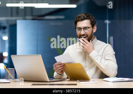 Ein glücklicher junger Mann sitzt im Büro an einem Laptop und liest einen erhaltenen Brief, freut sich über die gute Nachricht. Stockfoto