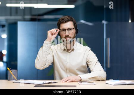 Porträt eines ernsthaften jungen Mannes, der im Büro am Schreibtisch in einem Headset vor der Kamera sitzt, auf die Kamera blickt und eine Brille hält. Stockfoto
