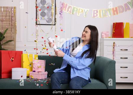 Glückliche Frau, die Konfetti-Popper auf dem Sofa zu Hause in die Luft jagt Stockfoto