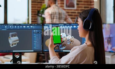 Asiatischer freiberuflicher Entwickler, der mit Kopfhörern und Chroma Key Screen arbeitet und einen neuen Prototyp für die Fertigungsindustrie entwickelt. Weibliche 3D-Modellierungsingenieurin in ihrem Heimbüro. Kamera B.. Stockfoto